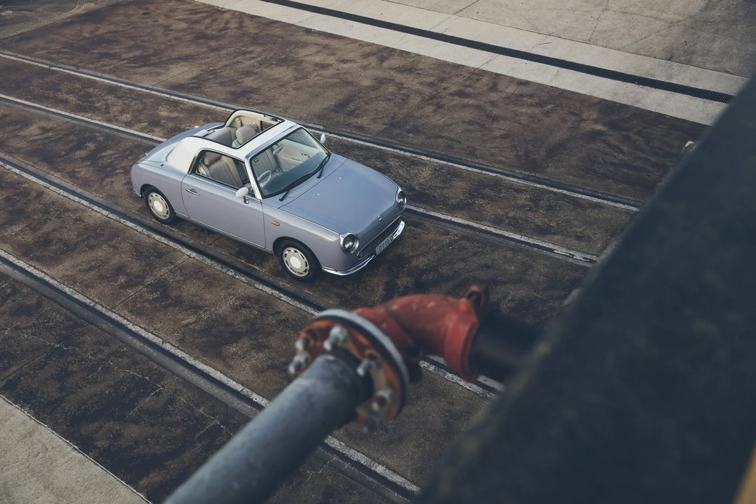 Pale blue Nissan Figaro seen from above at an industrial site, with a large pipe crossing the foreground.