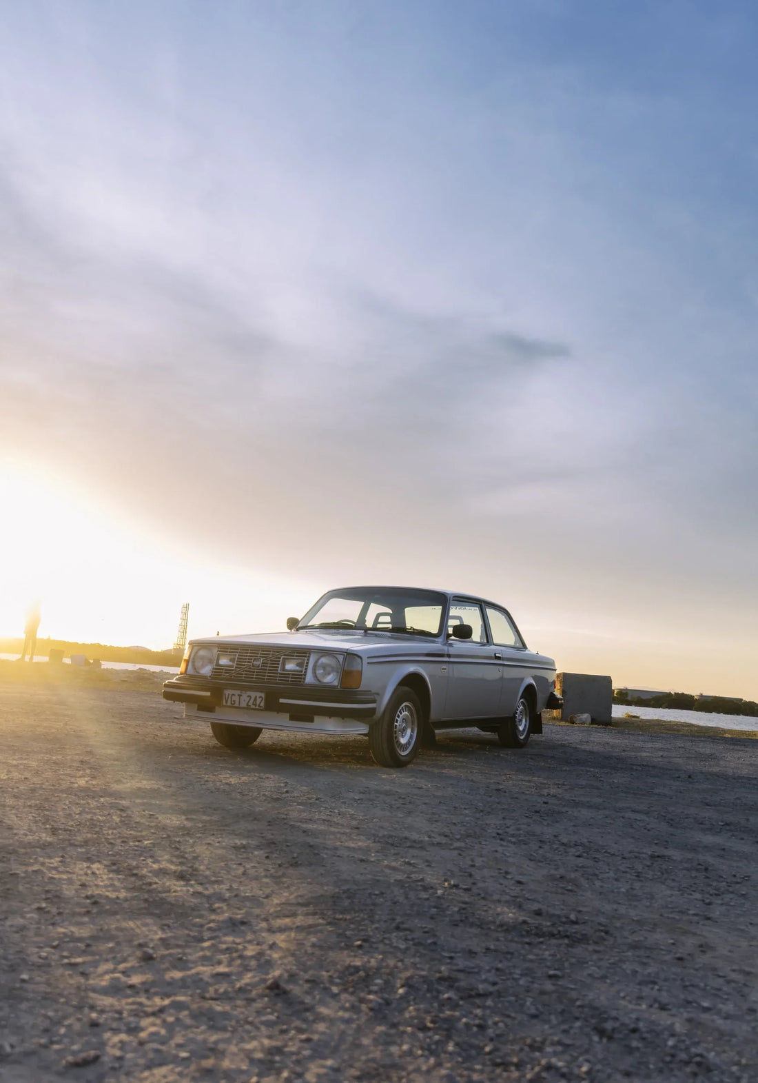 Silver Volvo 242 GT parked on gravel at sunset beneath a pale sky.