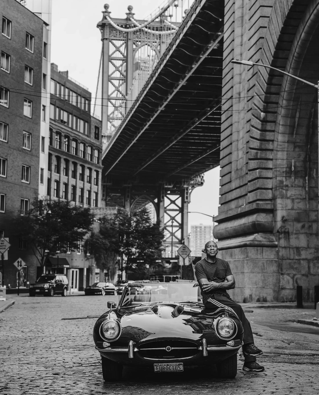 George Sweeper sitting on his 1968 Jaguar E-Type in black-and-white portrait beneath Manhattan Bridge in DUMBO, photograph by Shaun Maluga
