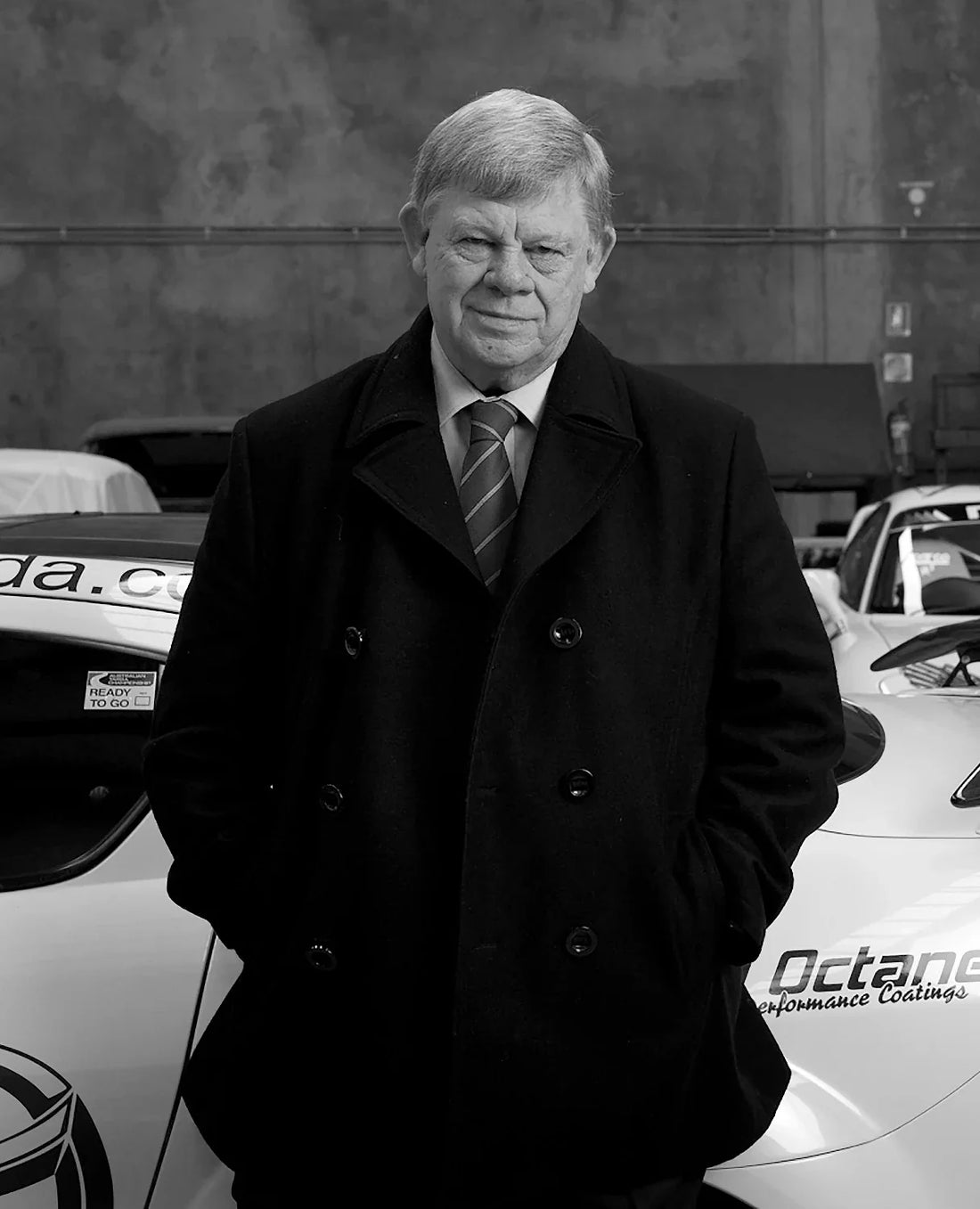 Black-and-white portrait of Allan Horsley standing in front of Mazda racing cars, photographed by Mazda Australia.