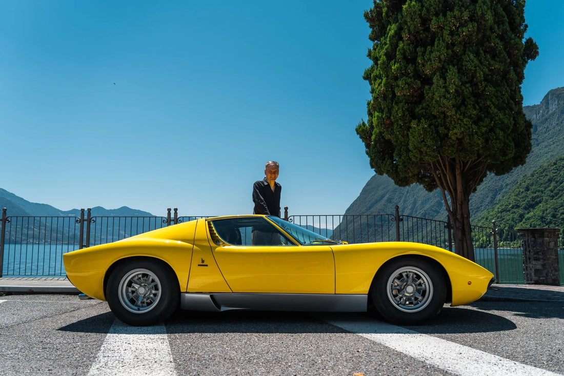 Side profile of a yellow Lamborghini Miura SV with Paolo Nespoli at a lakeside mountain overlook.
